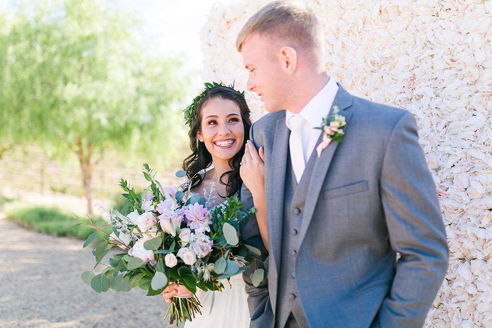 Temecula outdoor wedding at bel vino winery bride a line chiffon gown with straps and lace detail on bodice with a sweetheart neckline and white and green flower crown with groom grey notch lapel suit with a matching vest and white dress shirt with a long white tie and white and pink floral boutonniere hugging and holding hands bride holding white and green floral bridal bouquet