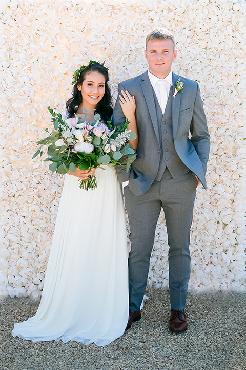 Temecula outdoor wedding at bel vino winery bride a line chiffon gown with straps and lace detail on bodice with a sweetheart neckline and white and green flower crown with groom grey notch lapel suit with a matching vest and white dress shirt with a long white tie and white and pink floral boutonniere holding hands and bride holding white and green floral bridal bouquet