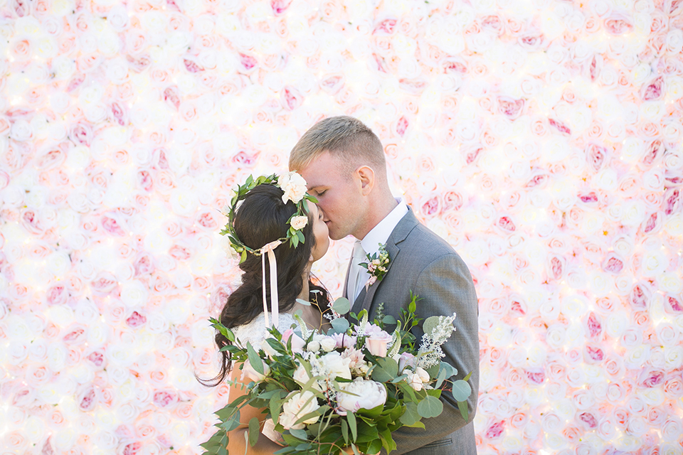 Temecula outdoor wedding at bel vino winery bride a line chiffon gown with straps and lace detail on bodice with a sweetheart neckline and white and green flower crown with groom grey notch lapel suit with a matching vest and white dress shirt with a long white tie and white and pink floral boutonniere kissing and bride holding white and green floral bridal bouquet