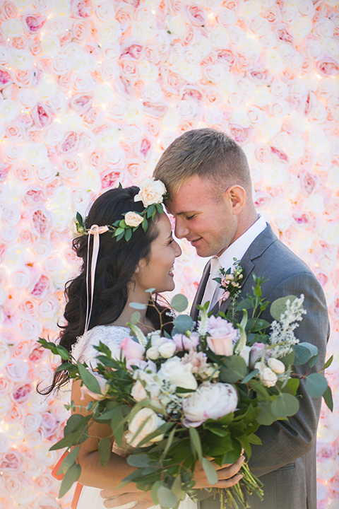 Temecula outdoor wedding at bel vino winery bride a line chiffon gown with straps and lace detail on bodice with a sweetheart neckline and white and green flower crown with groom grey notch lapel suit with a matching vest and white dress shirt with a long white tie and white and pink floral boutonniere hugging and bride holding white and green floral bridal bouquet