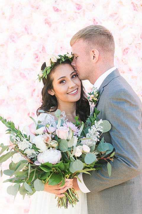 Temecula outdoor wedding at bel vino winery bride a line chiffon gown with straps and lace detail on bodice with a sweetheart neckline and white and green flower crown with groom grey notch lapel suit with a matching vest and white dress shirt with a long white tie and white and pink floral boutonniere hugging and bride holding white and green floral bridal bouquet