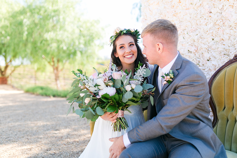 Temecula outdoor wedding at bel vino winery bride a line chiffon gown with straps and lace detail on bodice with a sweetheart neckline and white and green flower crown with groom grey notch lapel suit with a matching vest and white dress shirt with a long white tie and white and pink floral boutonniere sitting and bride holding white and green floral bridal bouquet