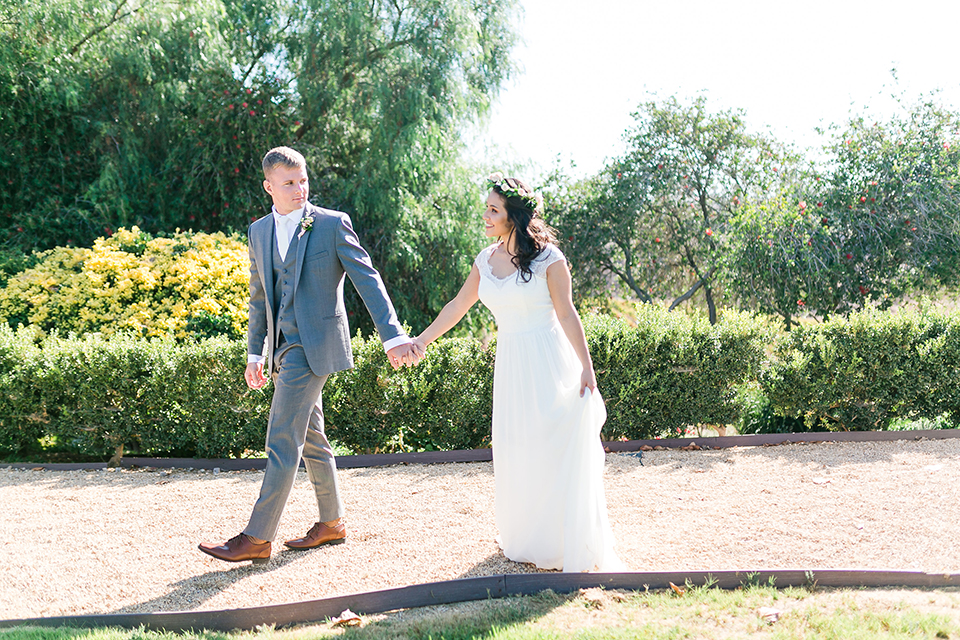 Temecula outdoor wedding at bel vino winery bride a line chiffon gown with straps and lace detail on bodice with a sweetheart neckline and white and green flower crown with groom grey notch lapel suit with a matching vest and white dress shirt with a long white tie and white and pink floral boutonniere holding hands and walking