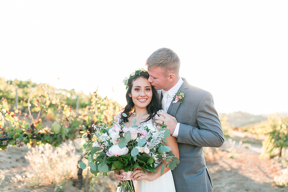 Temecula outdoor wedding at bel vino winery bride a line chiffon gown with straps and lace detail on bodice with a sweetheart neckline and white and green flower crown with groom grey notch lapel suit with a matching vest and white dress shirt with a long white tie and white and pink floral boutonniere hugging and kissing bride holding white and green floral bridal bouquet