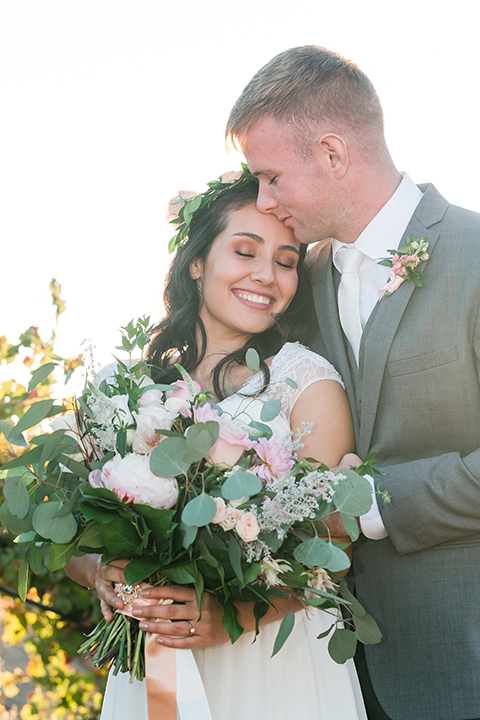 Temecula outdoor wedding at bel vino winery bride a line chiffon gown with straps and lace detail on bodice with a sweetheart neckline and white and green flower crown with groom grey notch lapel suit with a matching vest and white dress shirt with a long white tie and white and pink floral boutonniere hugging and kissing close up and bride holding white and green floral bridal bouquet