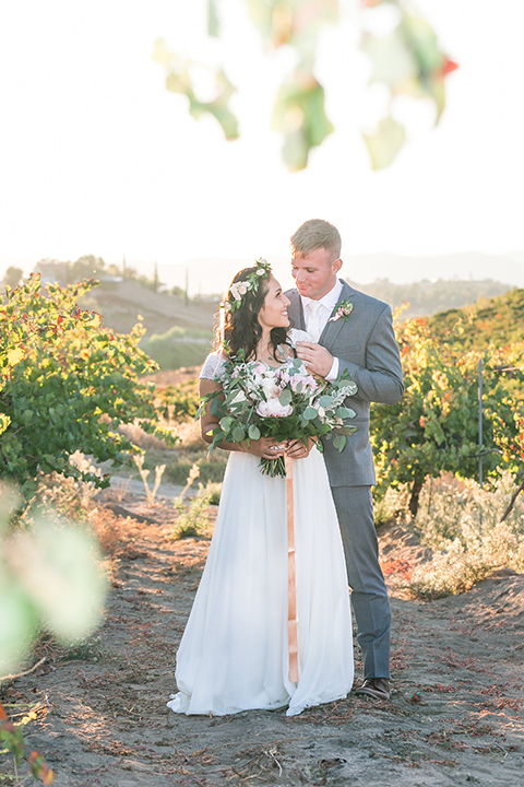 Temecula outdoor wedding at bel vino winery bride a line chiffon gown with straps and lace detail on bodice with a sweetheart neckline and white and green flower crown with groom grey notch lapel suit with a matching vest and white dress shirt with a long white tie and white and pink floral boutonniere hugging and smiling bride holding white and green floral bridal bouquet