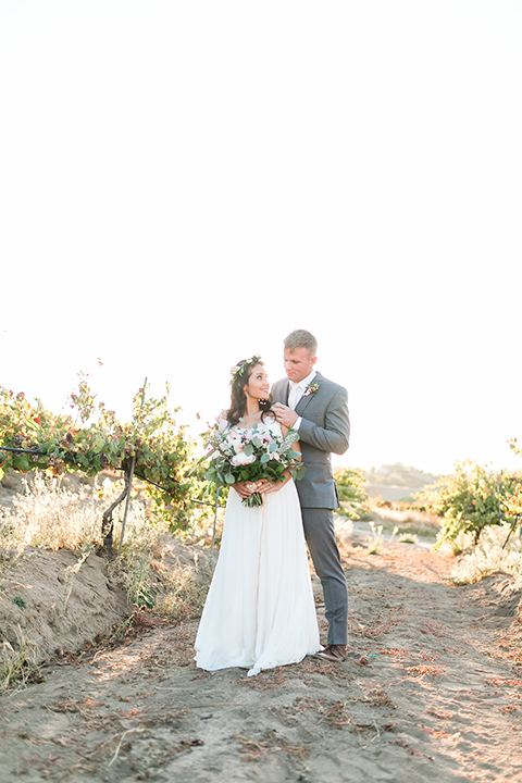 Temecula outdoor wedding at bel vino winery bride a line chiffon gown with straps and lace detail on bodice with a sweetheart neckline and white and green flower crown with groom grey notch lapel suit with a matching vest and white dress shirt with a long white tie and white and pink floral boutonniere standing and hugging bride holding white and green floral bridal bouquet