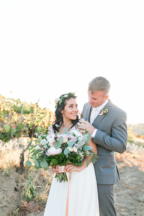 Temecula outdoor wedding at bel vino winery bride a line chiffon gown with straps and lace detail on bodice with a sweetheart neckline and white and green flower crown with groom grey notch lapel suit with a matching vest and white dress shirt with a long white tie and white and pink floral boutonniere hugging and bride holding white and green floral bridal bouquet