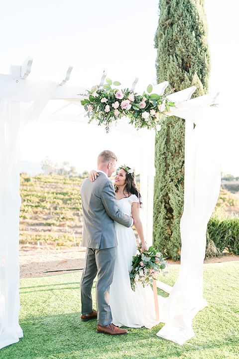 Temecula outdoor wedding at bel vino winery bride a line chiffon gown with straps and lace detail on bodice with a sweetheart neckline and white and green flower crown with groom grey notch lapel suit with a matching vest and white dress shirt with a long white tie and white and pink floral boutonniere hugging during ceremony and bride holding white and green floral bridal bouquet