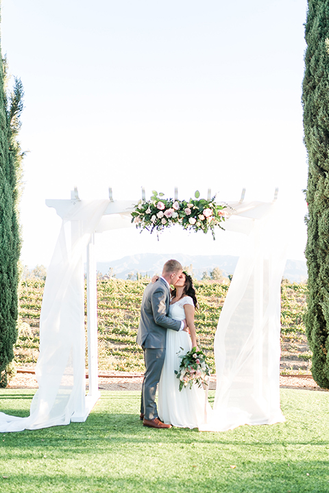 Temecula outdoor wedding at bel vino winery bride a line chiffon gown with straps and lace detail on bodice with a sweetheart neckline and white and green flower crown with groom grey notch lapel suit with a matching vest and white dress shirt with a long white tie and white and pink floral boutonniere kissing during ceremony and bride holding white and green floral bridal bouquet