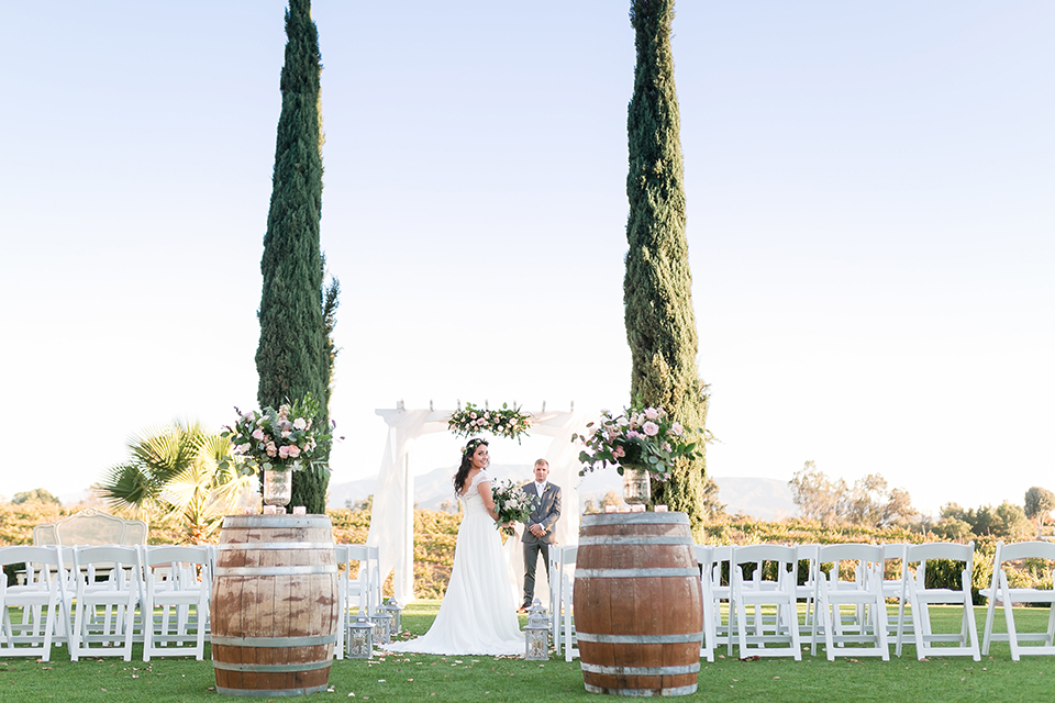 Temecula outdoor wedding at bel vino winery bride a line chiffon gown with straps and lace detail on bodice with a sweetheart neckline and white and green flower crown with groom grey notch lapel suit with a matching vest and white dress shirt with a long white tie and white and pink floral boutonniere standing during ceremony bride holding white and green floral bridal bouquet