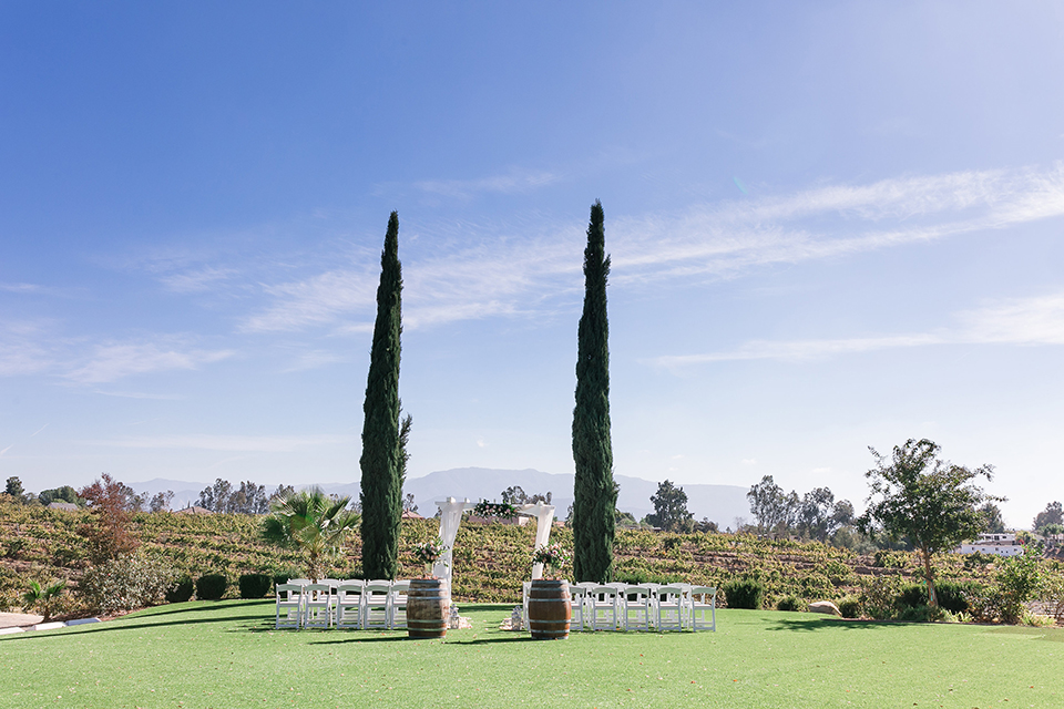 Temecula outdoor wedding at bel vino winery ceremony set up with white chairs and wine barrels with flower decor and white chiffon altar with white and green flower decor on top wedding photo idea for ceremony set up