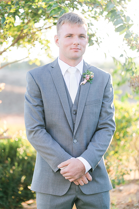 Temecula outdoor wedding at bel vino winery groom grey notch lapel suit with a matching vest and white dress shirt with a long white tie and white and pink floral boutonniere standing and crossing hands