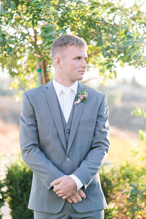 Temecula outdoor wedding at bel vino winery groom grey notch lapel suit with a matching vest and white dress shirt with a long white tie and white and pink floral boutonniere standing and crossing hands