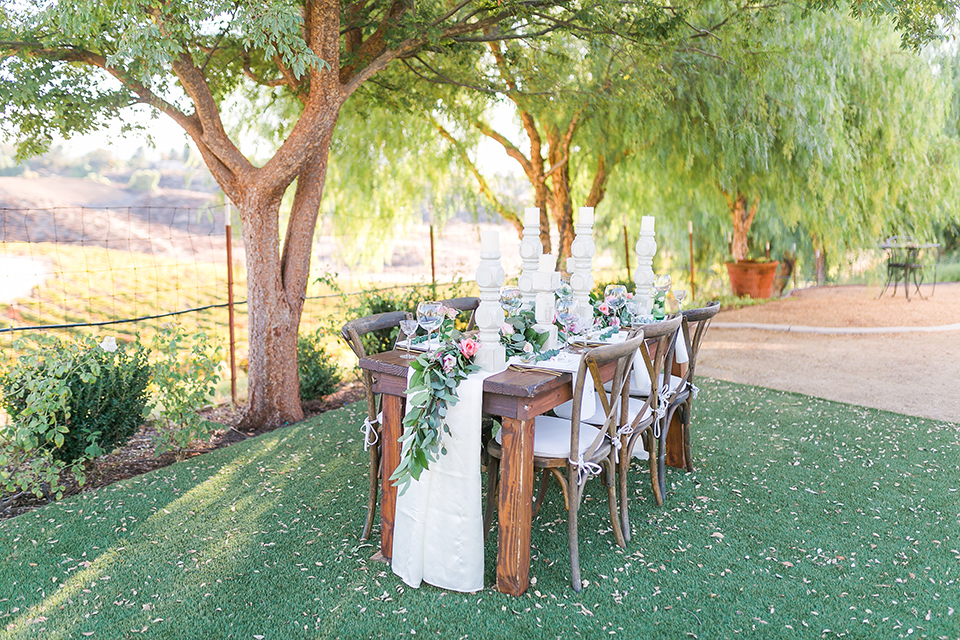 Temecula outdoor wedding at bel vino winery table set up dark brown wood table with white square place settings and pink and green garland flower decor in center and gold silverware decor with matching brown chairs and white table runner