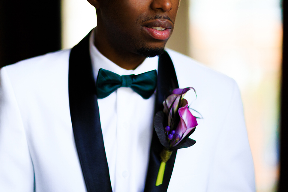 Boutonniere-style-white-shawl-lapel-tuxedo-with-a-dark-green-bow-tie-and-a-big-purple-floral-boutonniere