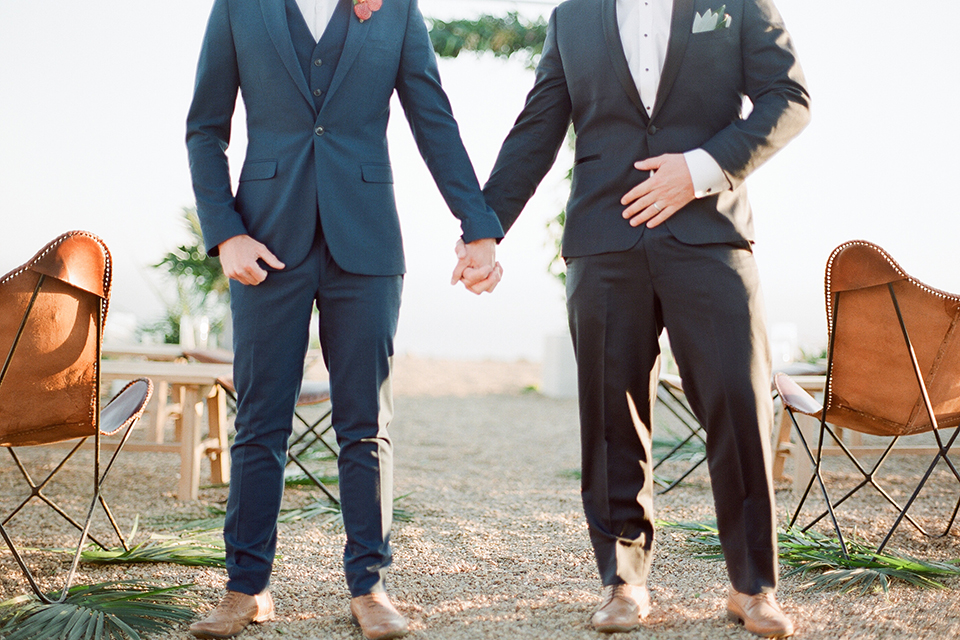 Los angeles same sex outdoor wedding at malibu solstice canyon groom navy shawl lapel tuxedo with a white dress shirt and black and white dot bow tie with groom blue notch lapel suit with a white dress shirt and white bow tie and orange floral boutonniere holding hands close up