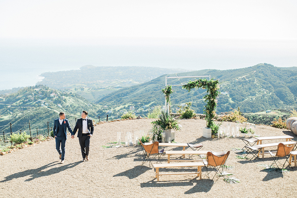 Los angeles same sex outdoor wedding at malibu solstice canyon groom navy shawl lapel tuxedo with a white dress shirt and black and white dot bow tie with groom blue notch lapel suit with a white dress shirt and white bow tie and orange floral boutonniere holding hands and walking by ceremony set up