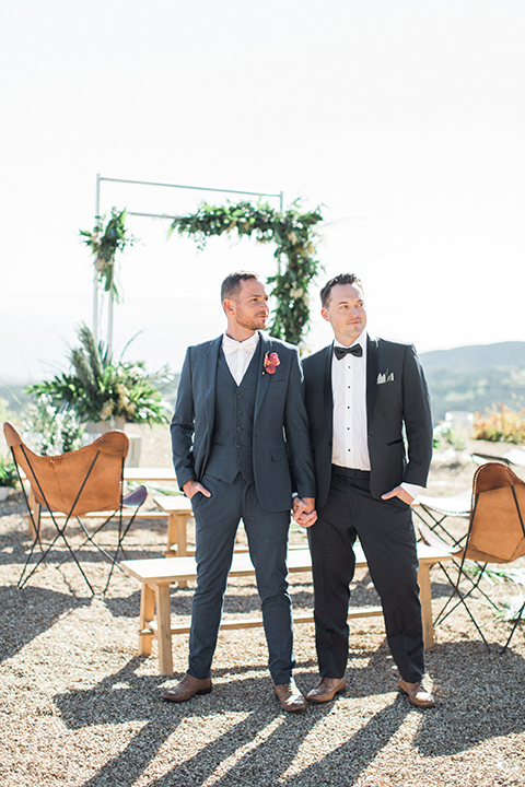 Los angeles same sex outdoor wedding at malibu solstice canyon groom navy shawl lapel tuxedo with a white dress shirt and black and white dot bow tie with groom blue notch lapel suit with a white dress shirt and white bow tie and orange floral boutonniere standing by ceremony set up holding hands