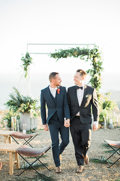 Los angeles same sex outdoor wedding at malibu solstice canyon groom navy shawl lapel tuxedo with a white dress shirt and black and white dot bow tie with groom blue notch lapel suit with a white dress shirt and white bow tie and orange floral boutonniere holding hands walking down aisle for ceremony