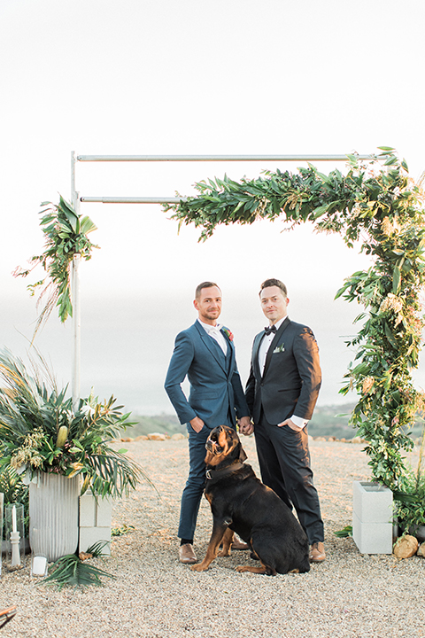 Los angeles same sex outdoor wedding at malibu solstice canyon groom navy shawl lapel tuxedo with a white dress shirt and black and white dot bow tie with groom blue notch lapel suit with a white dress shirt and white bow tie and orange floral boutonniere hugging and holding hands standing with dog at ceremony set up