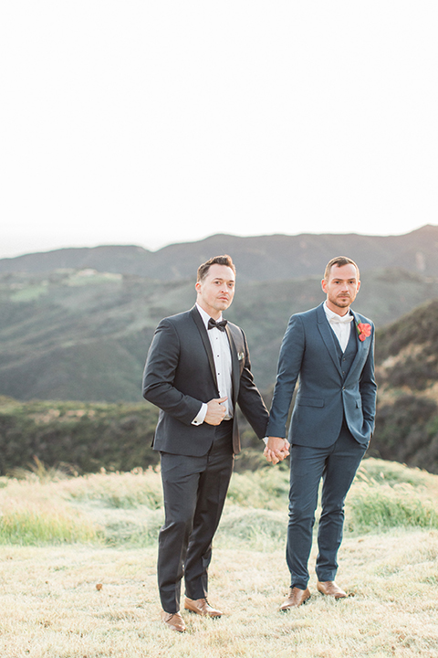 Los angeles same sex outdoor wedding at malibu solstice canyon groom navy shawl lapel tuxedo with a white dress shirt and black and white dot bow tie with groom blue notch lapel suit with a white dress shirt and white bow tie and orange floral boutonniere holding hands standing