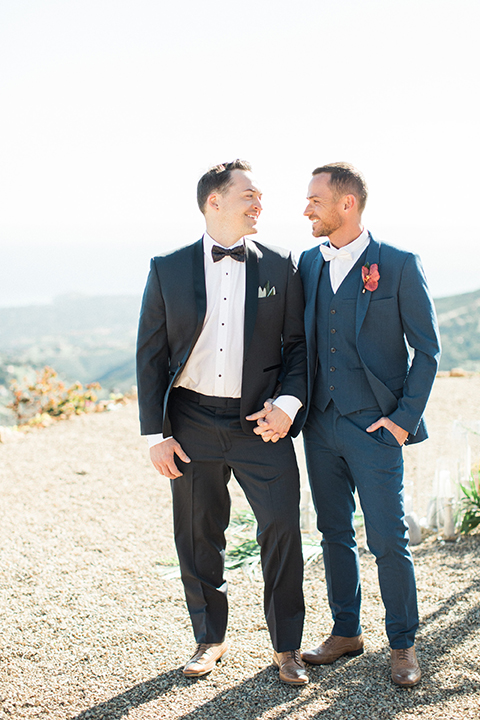 Los angeles same sex outdoor wedding at malibu solstice canyon groom navy shawl lapel tuxedo with a white dress shirt and black and white dot bow tie with groom blue notch lapel suit with a white dress shirt and white bow tie and orange floral boutonniere hugging and holding hands