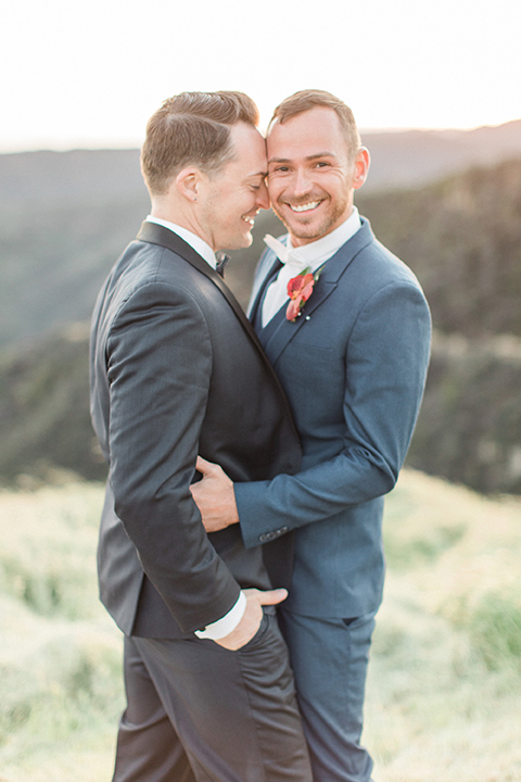 Los angeles same sex outdoor wedding at malibu solstice canyon groom navy shawl lapel tuxedo with a white dress shirt and black and white dot bow tie with groom blue notch lapel suit with a white dress shirt and white bow tie and orange floral boutonniere hugging and smiling close up