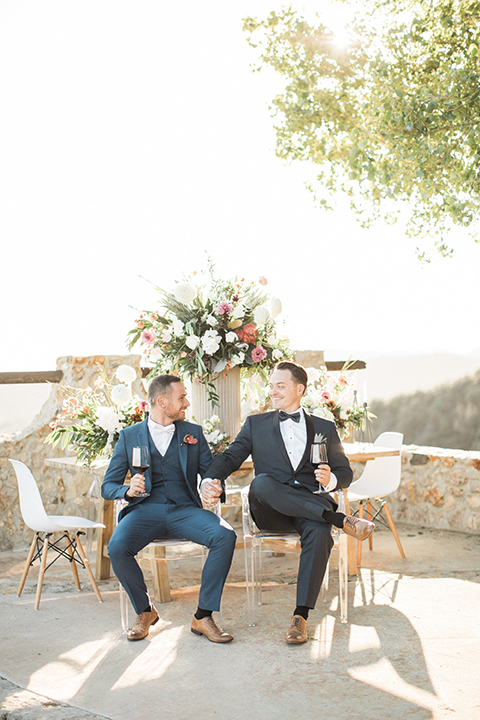 Los angeles same sex outdoor wedding at malibu solstice canyon groom navy shawl lapel tuxedo with a white dress shirt and black and white dot bow tie with groom blue notch lapel suit with a white dress shirt and white bow tie and orange floral boutonniere sitting at table holding wine smiling