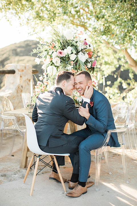 Los angeles same sex outdoor wedding at malibu solstice canyon groom navy shawl lapel tuxedo with a white dress shirt and black and white dot bow tie with groom blue notch lapel suit with a white dress shirt and white bow tie and orange floral boutonniere sitting at table hugging