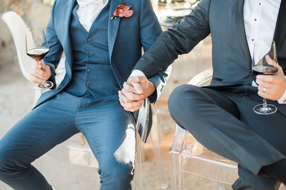 Los angeles same sex outdoor wedding at malibu solstice canyon groom navy shawl lapel tuxedo with a white dress shirt and black and white dot bow tie with groom blue notch lapel suit with a white dress shirt and white bow tie and orange floral boutonniere sitting at table holding hands close up on hands front view