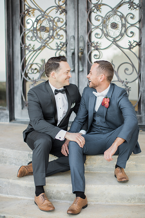 Los angeles same sex outdoor wedding at malibu solstice canyon groom navy shawl lapel tuxedo with a white dress shirt and black and white dot bow tie with groom blue notch lapel suit with a white dress shirt and white bow tie and orange floral boutonniere sitting on steps holding hands front view