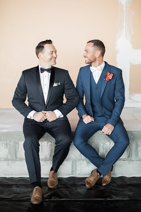 Los angeles same sex outdoor wedding at malibu solstice canyon groom navy shawl lapel tuxedo with a white dress shirt and black and white dot bow tie with groom blue notch lapel suit with a white dress shirt and white bow tie and orange floral boutonniere sitting and smiling front view