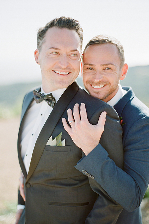 Los angeles same sex outdoor wedding at malibu solstice canyon groom navy shawl lapel tuxedo with a white dress shirt and black and white dot bow tie with groom blue notch lapel suit with a white dress shirt and white bow tie and orange floral boutonniere hugging close up front view