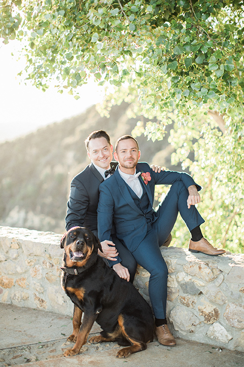 Los angeles same sex outdoor wedding at malibu solstice canyon groom navy shawl lapel tuxedo with a white dress shirt and black and white dot bow tie with groom blue notch lapel suit with a white dress shirt and white bow tie and orange floral boutonniere sitting on wall with dog front view