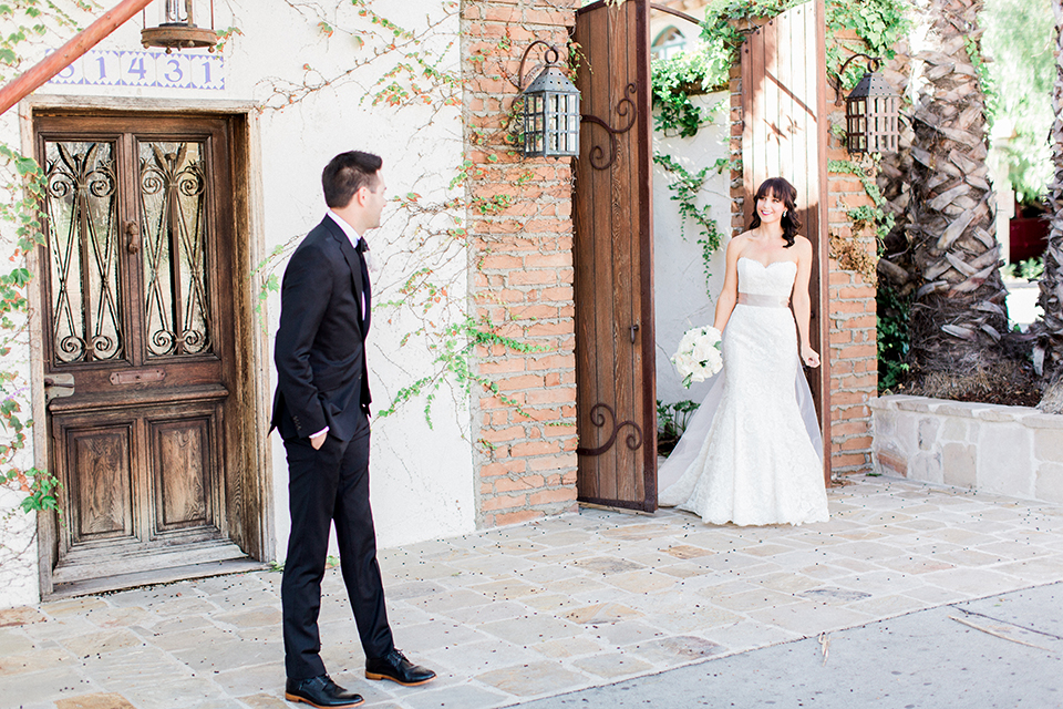 Fall wedding at the villa san juan capistrano bride strapless mermaid fit gown with a sweetheart neckline and ribbon belt with bow in the back with groom black notch lapel suit with a white dress shirt and black bow tie with a black and white plaid pocket square and white floral boutonniere first look bride and groom smiling and walking