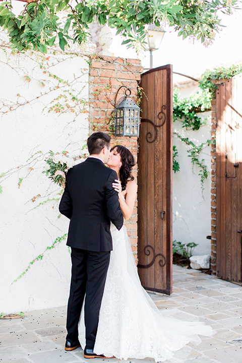 Fall wedding at the villa san juan capistrano bride strapless mermaid fit gown with a sweetheart neckline and ribbon belt with bow in the back with groom black notch lapel suit with a white dress shirt and black bow tie with a black and white plaid pocket square and white floral boutonniere first look bride and groom kissing bride holding white floral bridal bouquet