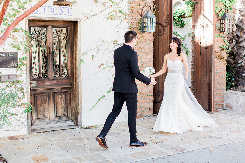 Fall wedding at the villa san juan capistrano bride strapless mermaid fit gown with a sweetheart neckline and ribbon belt with bow in the back with groom black notch lapel suit with a white dress shirt and black bow tie with a black and white plaid pocket square and white floral boutonniere first look bride and groom walking and smiling bride holding white floral bridal bouquet