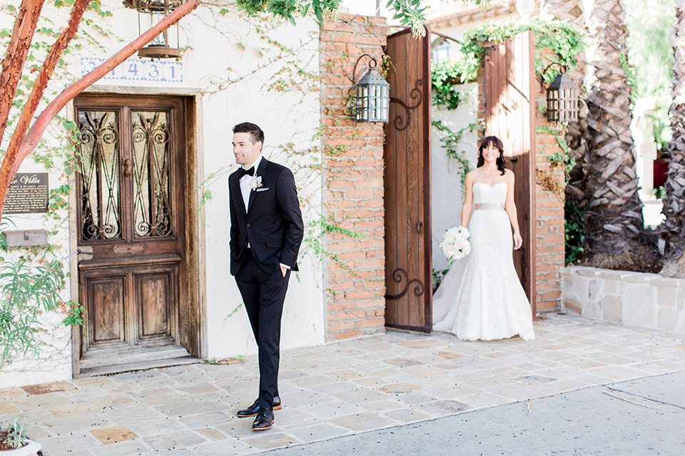Fall wedding at the villa san juan capistrano bride strapless mermaid fit gown with a sweetheart neckline and ribbon belt with bow in the back with groom black notch lapel suit with a white dress shirt and black bow tie with a black and white plaid pocket square and white floral boutonniere first look bride walking behind groom
