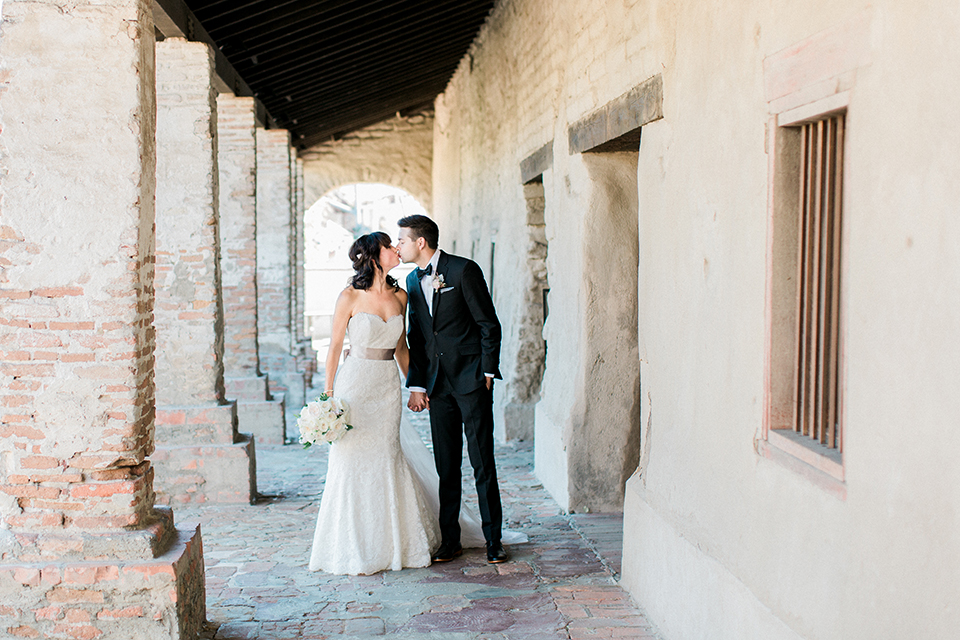 Fall wedding at the villa san juan capistrano bride strapless mermaid fit gown with a sweetheart neckline and ribbon belt with bow in the back with groom black notch lapel suit with a white dress shirt and black bow tie with a black and white plaid pocket square and white floral boutonniere holding hands and kissing bride holding white floral bridal bouquet