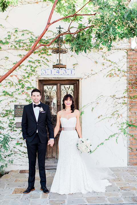 Fall wedding at the villa san juan capistrano bride strapless mermaid fit gown with a sweetheart neckline and ribbon belt with bow in the back with groom black notch lapel suit with a white dress shirt and black bow tie with a black and white plaid pocket square and white floral boutonniere holding hands and smiling bride holding white floral bridal bouquet