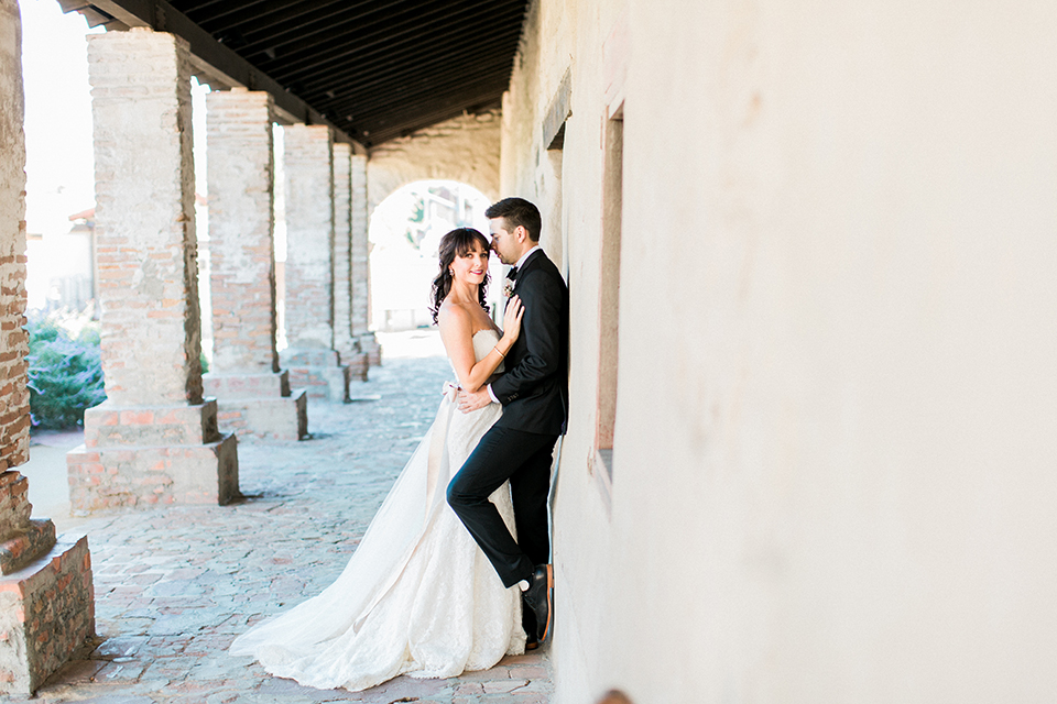 Fall wedding at the villa san juan capistrano bride strapless mermaid fit gown with a sweetheart neckline and ribbon belt with bow in the back with groom black notch lapel suit with a white dress shirt and black bow tie with a black and white plaid pocket square and white floral boutonniere standing against wall and hugging