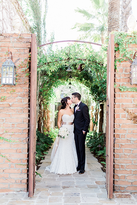 Fall wedding at the villa san juan capistrano bride strapless mermaid fit gown with a sweetheart neckline and ribbon belt with bow in the back with groom black notch lapel suit with a white dress shirt and black bow tie with a black and white plaid pocket square and white floral boutonniere standing and kissing bride holding white floral bridal bouquet