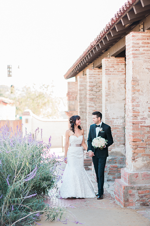 Fall wedding at the villa san juan capistrano bride strapless mermaid fit gown with a sweetheart neckline and ribbon belt with bow in the back with groom black notch lapel suit with a white dress shirt and black bow tie with a black and white plaid pocket square and white floral boutonniere standing and holding hands looking at each other smiling groom holding white floral bridal bouquet