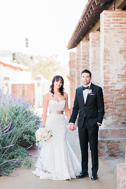 Fall wedding at the villa san juan capistrano bride strapless mermaid fit gown with a sweetheart neckline and ribbon belt with bow in the back with groom black notch lapel suit with a white dress shirt and black bow tie with a black and white plaid pocket square and white floral boutonniere standing and holding hands front view bride holding white floral bridal bouquet