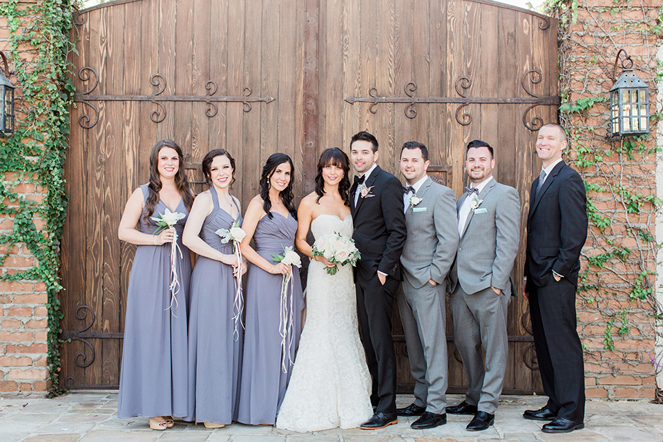 Fall wedding at the villa san juan capistrano bride strapless mermaid fit gown with a sweetheart neckline and ribbon belt with bow in the back with groom black notch lapel suit with a white dress shirt and black bow tie with a black and white plaid pocket square and white floral boutonniere standing with wedding party bridesmaids long dusty blue dresses with white floral bouquets with ribbon decor and groomsmen grey notch lapel suits with matching grey striped bow ties