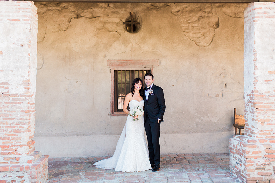 Fall wedding at the villa san juan capistrano bride strapless mermaid fit gown with a sweetheart neckline and ribbon belt with bow in the back with groom black notch lapel suit with a white dress shirt and black bow tie with a black and white plaid pocket square and white floral boutonniere 