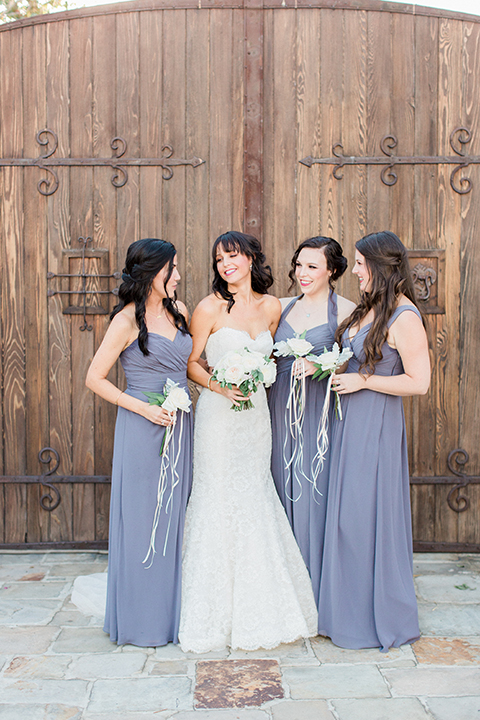 Fall wedding at the villa san juan capistrano bride strapless mermaid fit gown with a sweetheart neckline and ribbon belt with bow in the back holding white floral bridal bouquet standing with bridesmaids smiling wearing dusty blue dresses and holding white floral bouquets