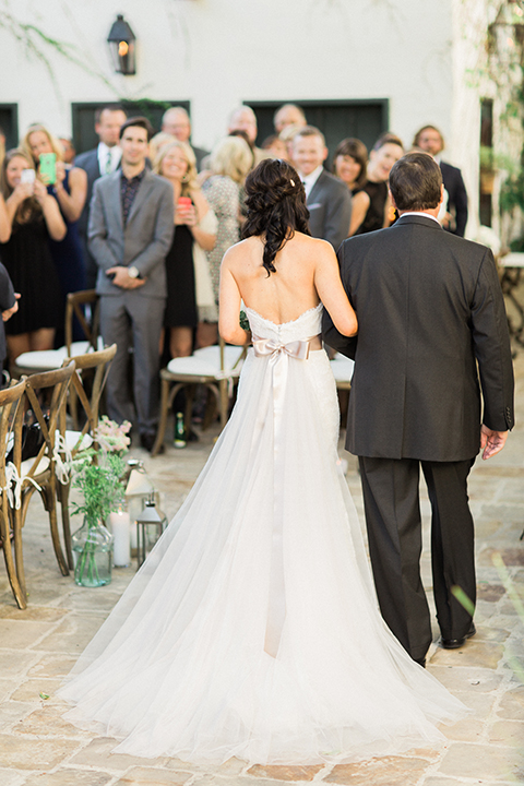 Fall wedding at the villa san juan capistrano bride strapless mermaid fit gown with a sweetheart neckline and ribbon belt with bow in the back holding white floral bridal bouquet walking down the aisle with dad back view
