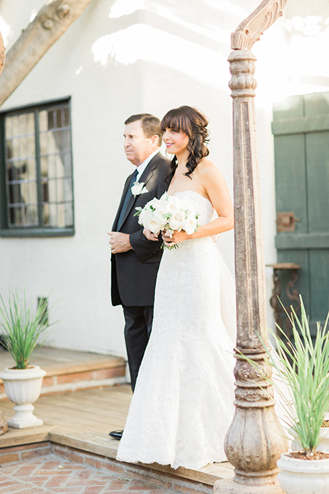 Fall wedding at the villa san juan capistrano bride strapless mermaid fit gown with a sweetheart neckline and ribbon belt with bow in the back holding white floral bridal bouquet walking down the aisle with dad smiling